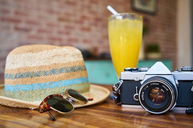 Summer essentials on a wooden table: straw hat, sunglasses, vintage camera, and a refreshing summer drink in a glass.