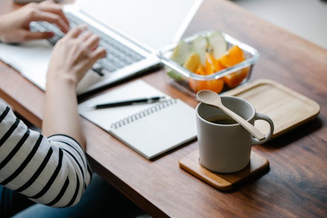 Person typing on a laptop with a notebook, pen, cup of tea, and a bowl of fresh fruit on a wooden table, representing a calm voice and mindful workspace.